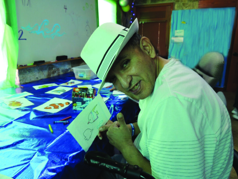 Older man giving thumbs up while sitting in a chair holding a drawing of fish. He is smiling and wearing a white hat.