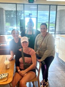 Therapist team Meredith Simone and Kendall Peterson with patient Ms. Timani at starbucks. Patient is sitting down at a table with a drink and both therapists are standing behind her smiling. Starbucks employee is also standing behind them smiling.