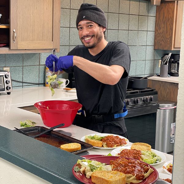 Patient Brandyn helping prep food in the residential program kitchen. He is breaking up lettuce in his hands and smiling.
