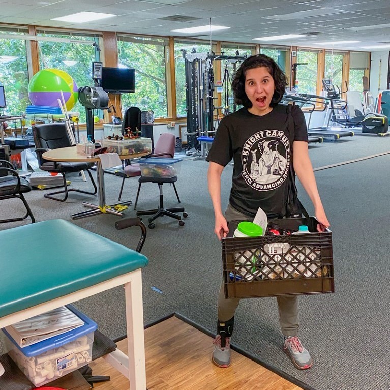 Parisa is holding a milk crate with items inside while she stands with a foot brace on. She is standing in a therapy gym.