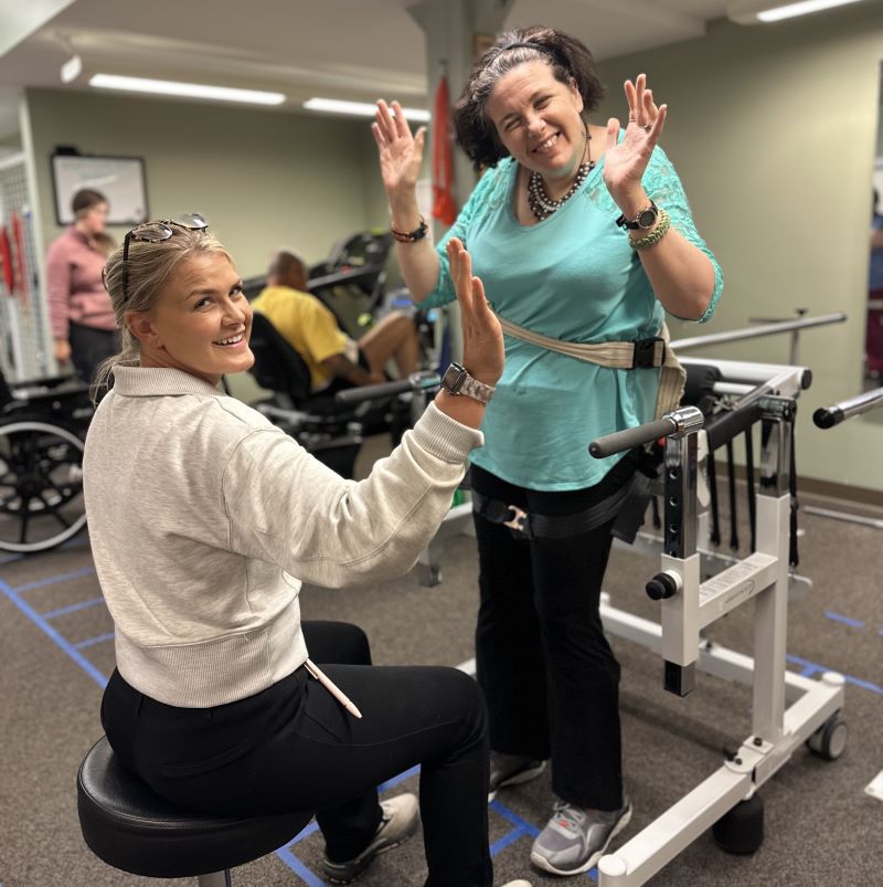 Patient Darci is standing with the help of the Neurogym Bungee Trainer which has strapped to the system. She is smiling with her hands in the air. Her therapist is sitting in front of her smiling.