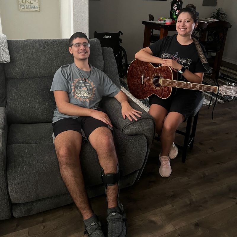 Gavin is sitting on the couch with his leg brace on, while his music therapist is sitting besides him in a chair holding a guitar. Both are smiling.