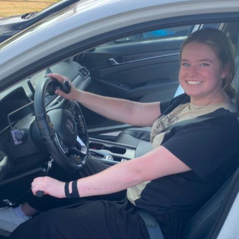 Patient Liz is sitting in a car with adaptive controls. She is smiling.