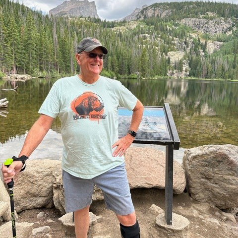 Bill is smiling while wearing a hat and sunglasses. He is hiking and holding his hiking pole. He is standing outdoors in front of a lake and trees and mountains.